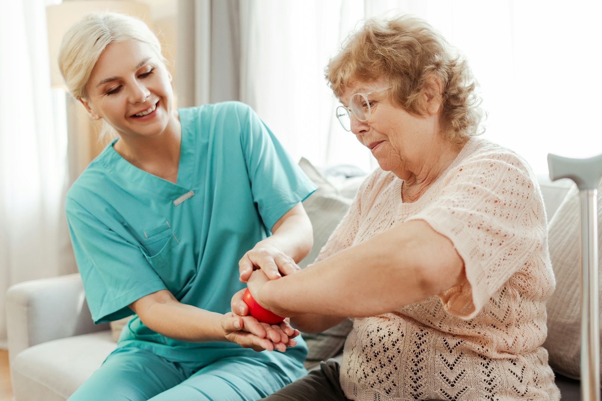 Smiling nurse assisting elderly woman exercising with red ball at home